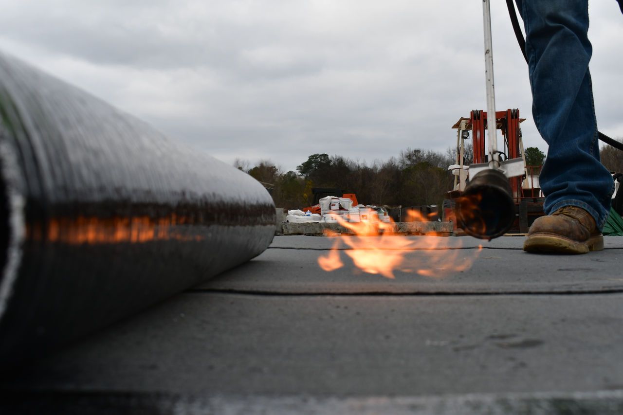 A worker using a torch flame to apply a roll of dark roofing membrane on an outdoor job site.
